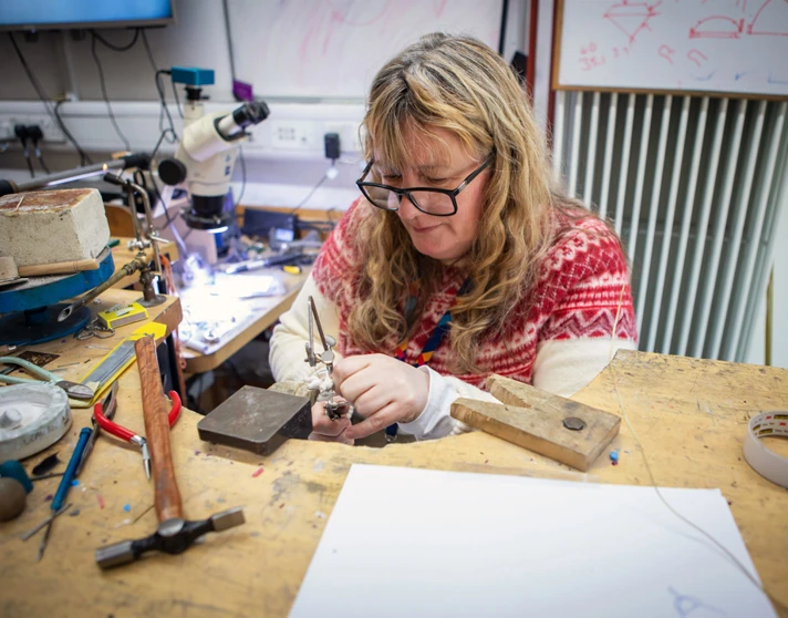 A female jewellery lecturer with long blonde hair and glasses, wearing a red and white sweater, working at a jewellery-making bench in a workshop filled with tools and equipment. A female jewellery lecturer with long blonde hair and glasses, wearing a red and white sweater, working at a jewellery-making bench in a workshop filled with tools and equipment.